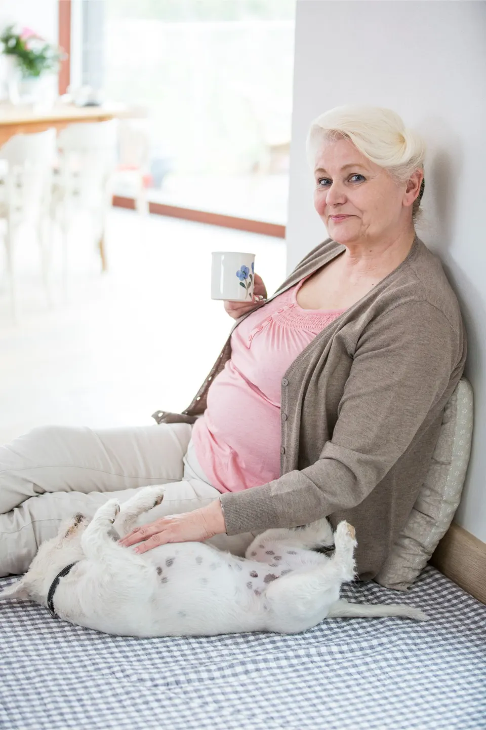 Femme âgée assise par terre, une tasse à la main, caressant le ventre d'un petit chien blanc couché sur le dos