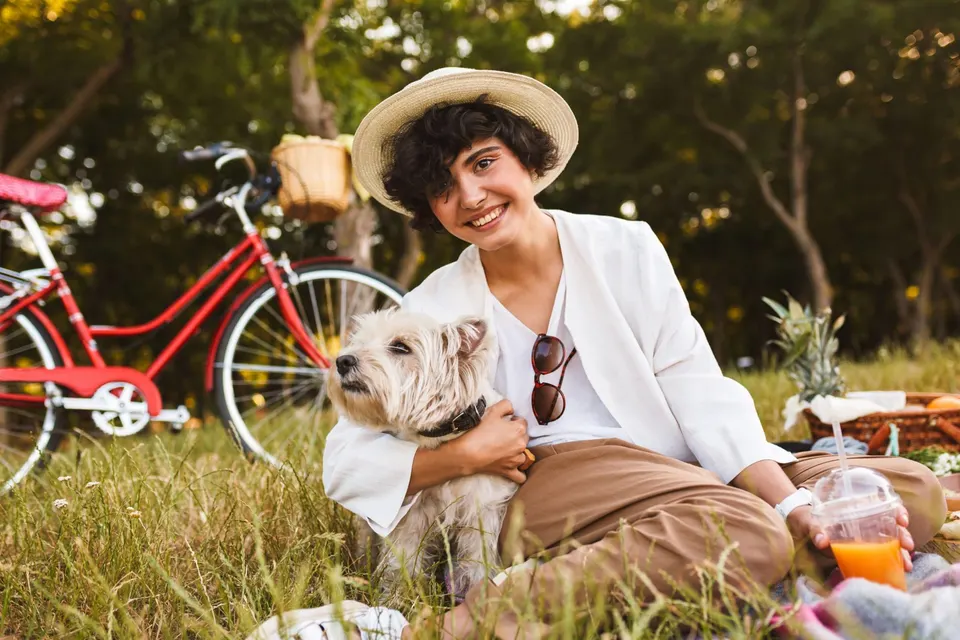 Une jeune femme au chapeau de paille serre un westie contre elle lors d'un pique-nique dans l'herbe, un vélo rouge derrière eux